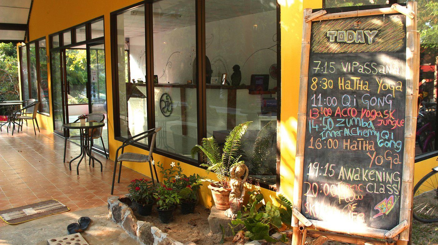 Yoga studio entrance with sign; yellow exterior, glass windows, plants, chairs and tables on a porch.