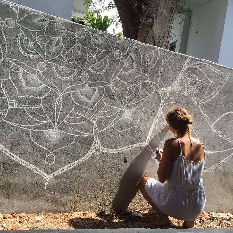 Woman in white dress drawing mandala on gray concrete wall outdoors.