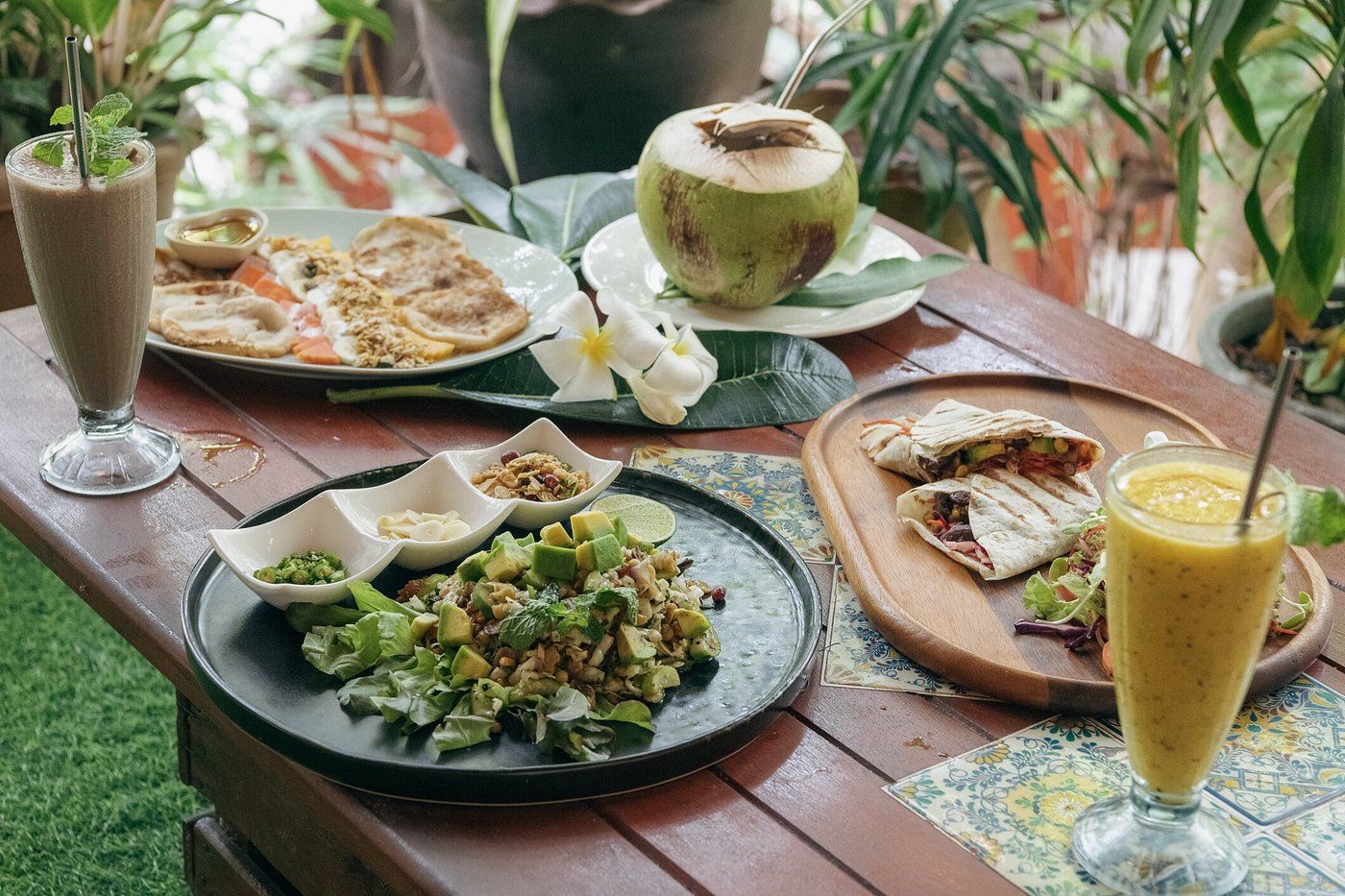 Healthy food spread on a table with smoothies, coconut, and a green salad in a tropical setting.
