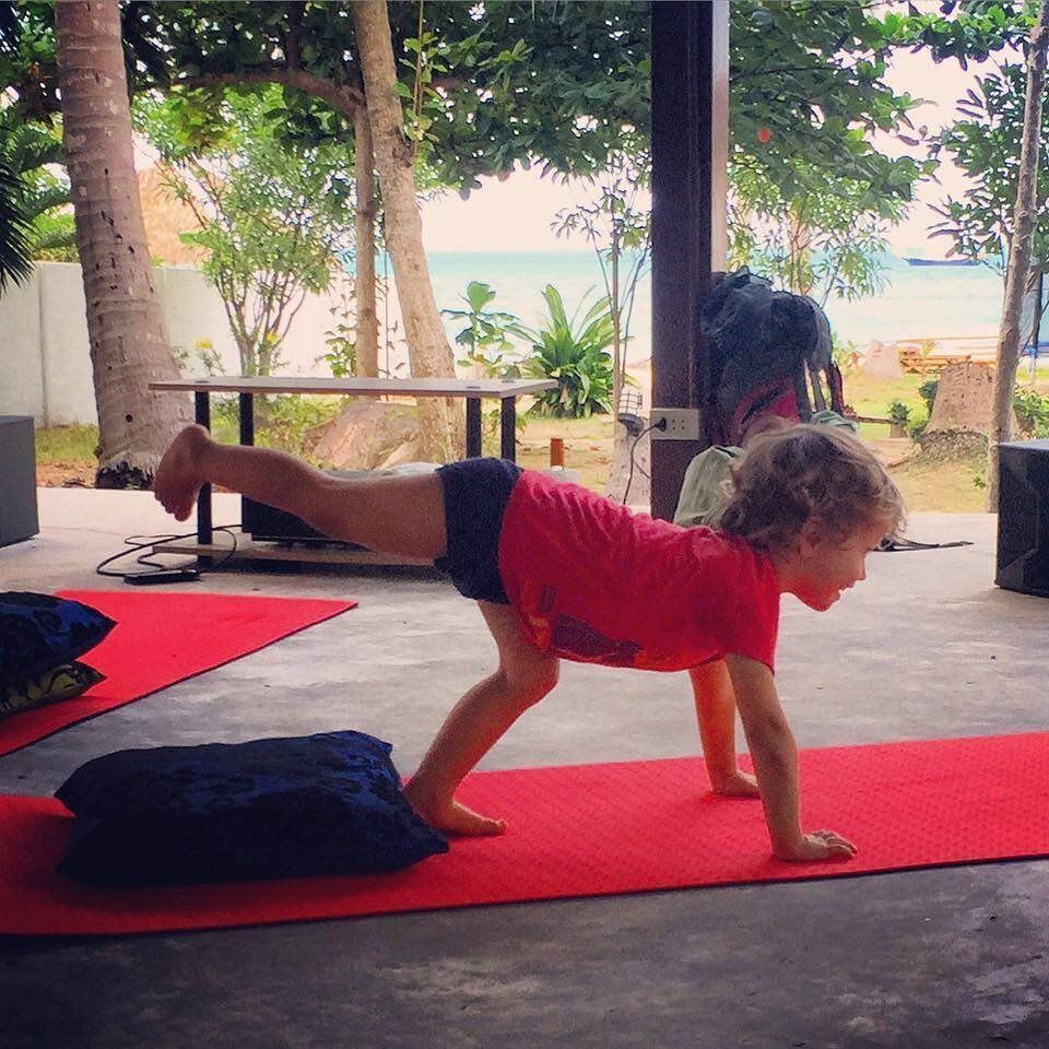 Young child doing yoga pose on a red mat, leg extended, outside with ocean view.
