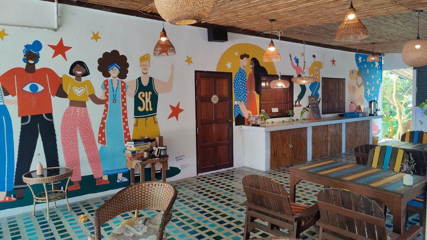 Colorful cafe interior with a mural of diverse figures. Wicker furniture, tiled floor, and hanging lights.