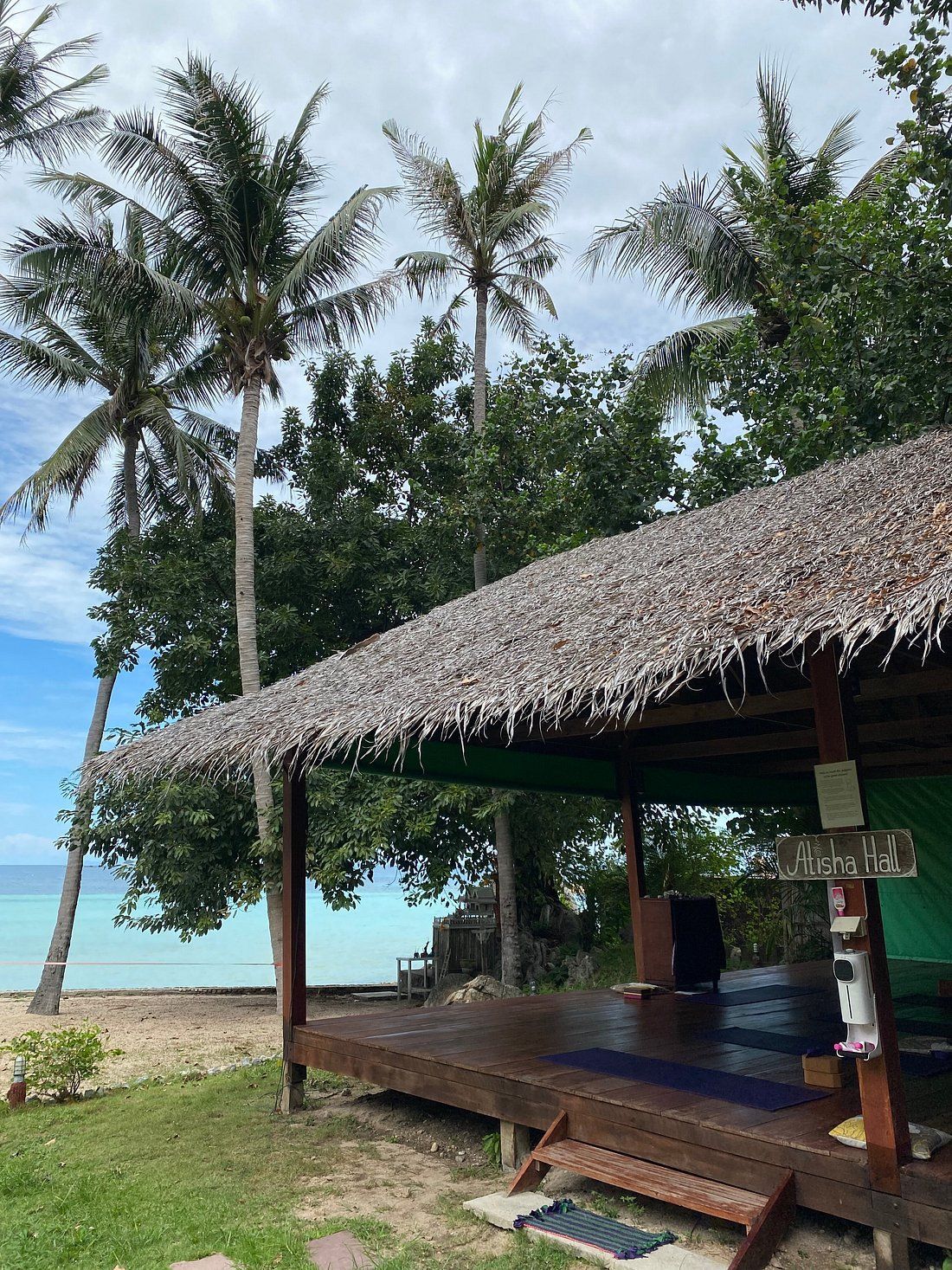 Thatched roof hut with a wooden deck, palm trees, and the ocean in the background.