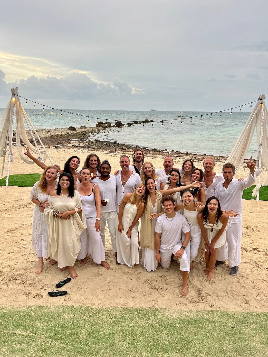 Group of people in white clothing on a beach with cloudy sky and ocean, posing and smiling.