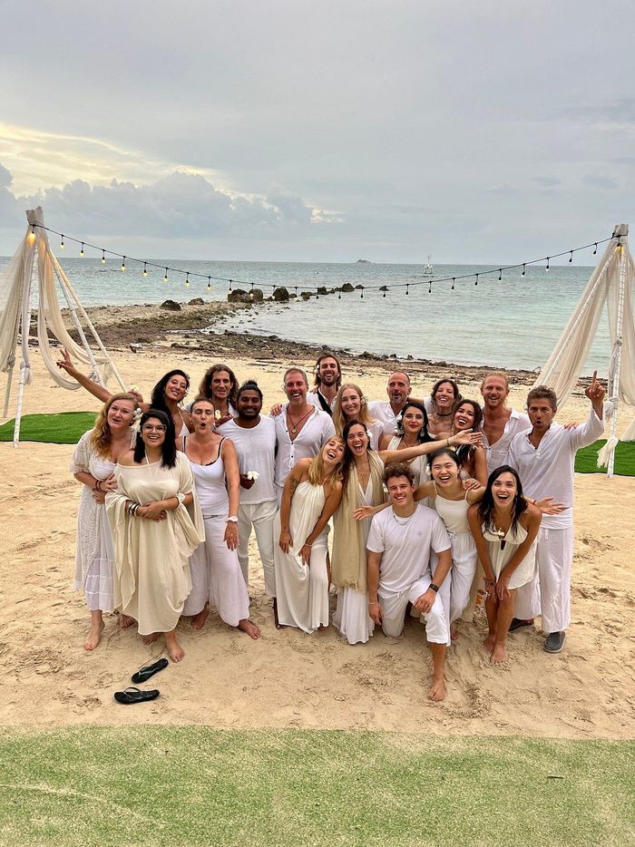 Group of people in white clothing on a beach with cloudy sky and ocean, posing.