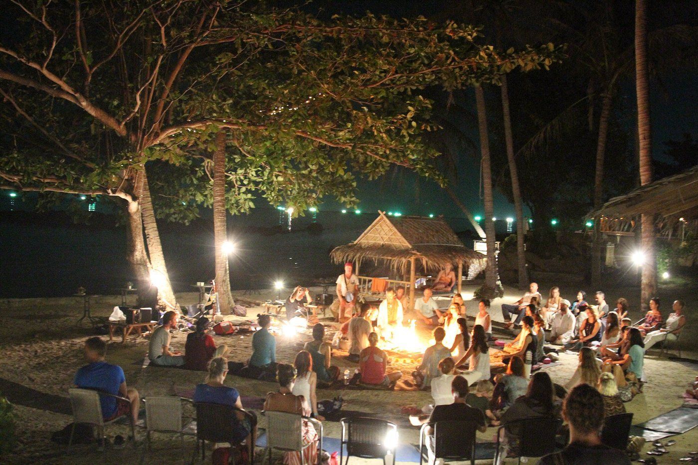 People gathered around a bonfire on a beach at night. Palm trees and lights illuminate the scene.
