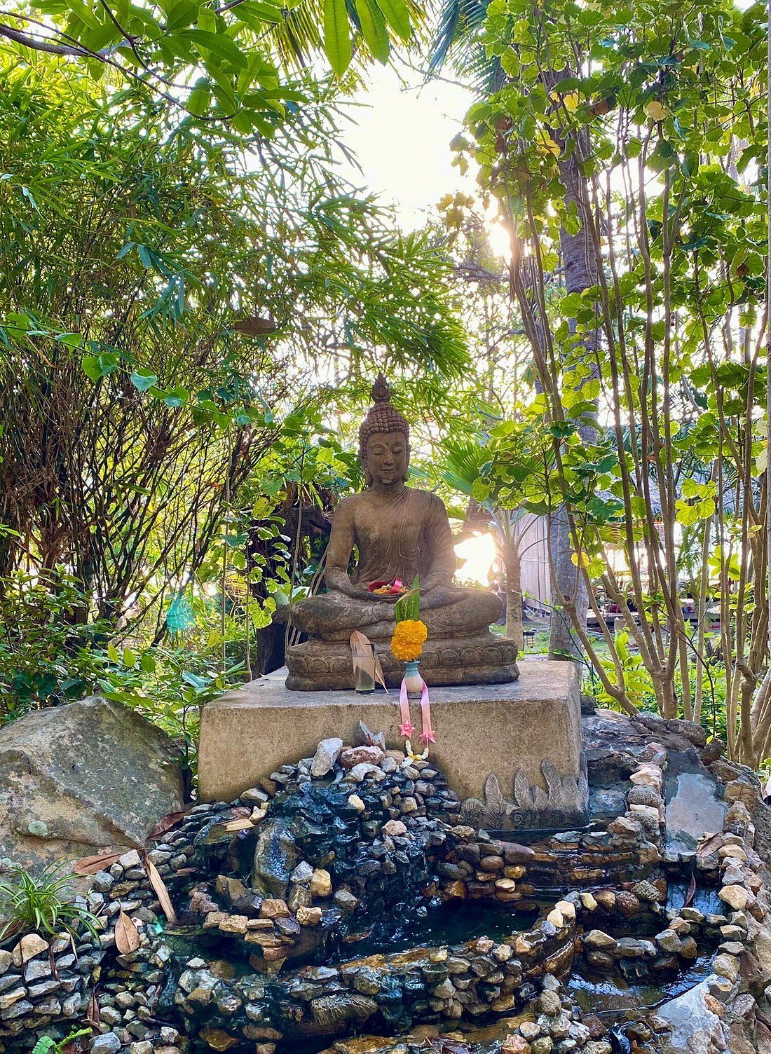 Stone Buddha statue seated on a platform, surrounded by greenery. Sunlight shines through trees.