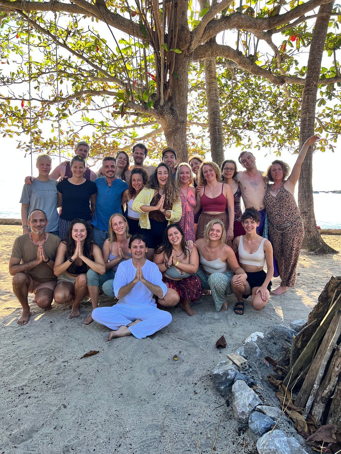 Group of people smiling, posing on a beach under a tree.