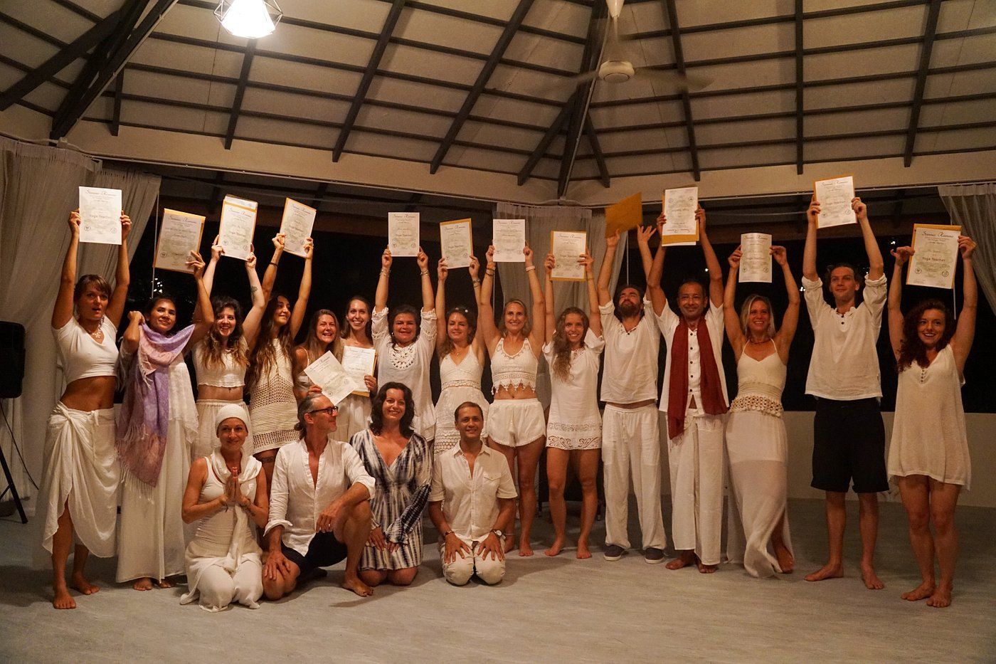Group of people in white clothing holding up certificates; indoors, under a wooden ceiling.