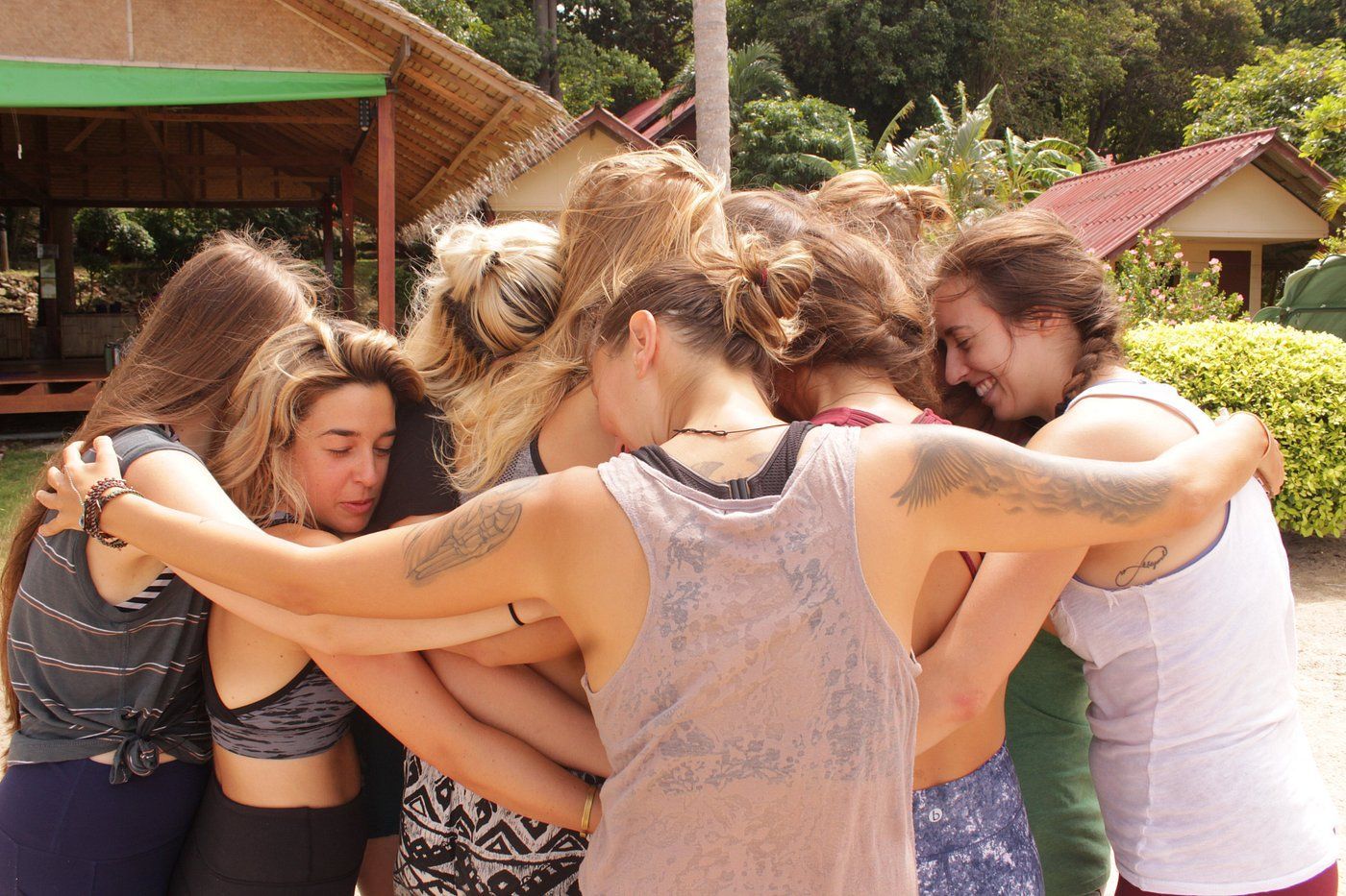 Group of women in a tight embrace outdoors, sunlit with visible tattoos and varying hair colors.