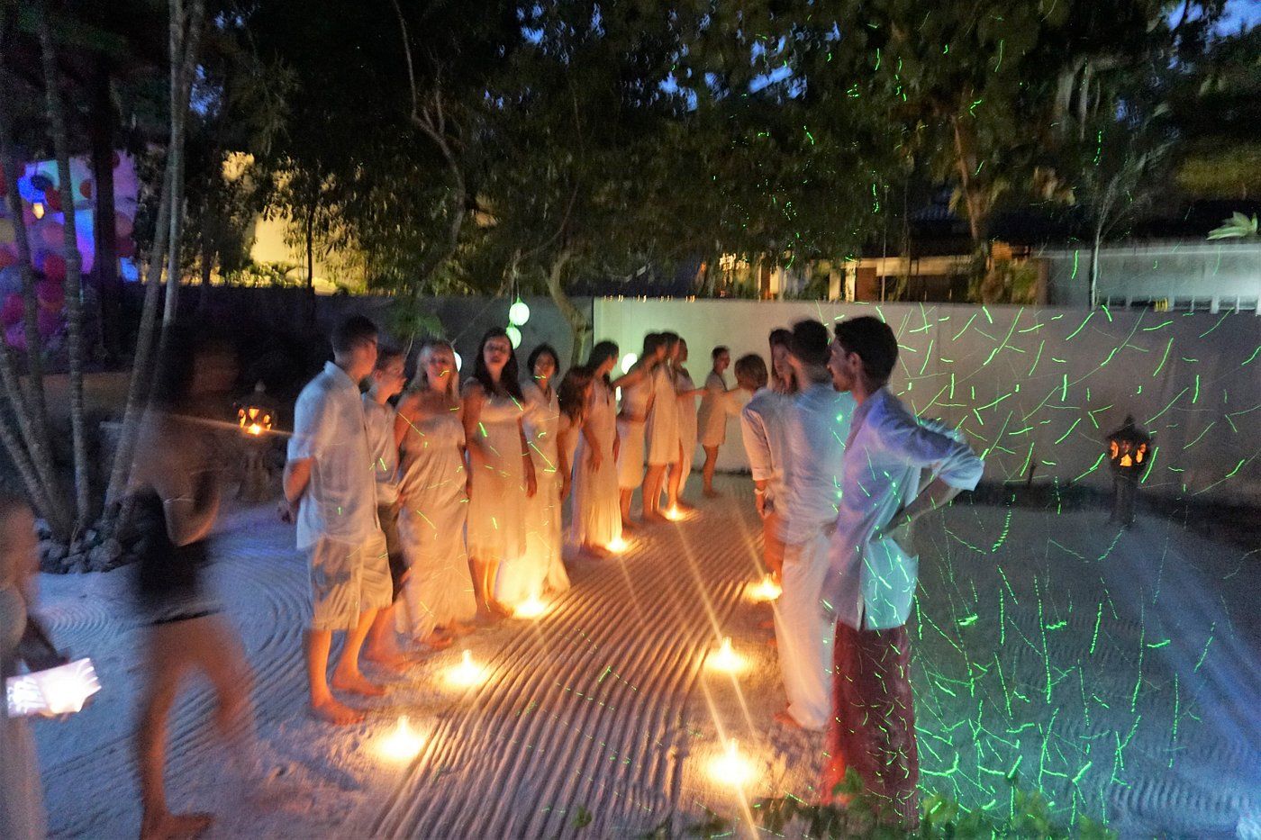 People in white clothing standing in a row, lit by candles and disco lights outside at night.