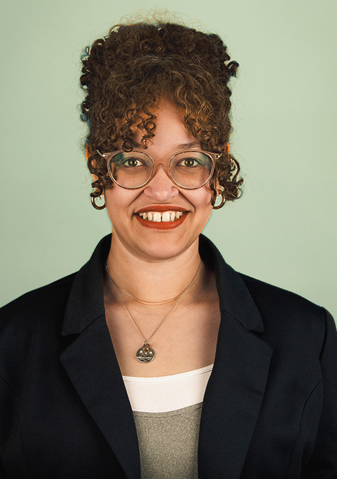 Jacqui Apollos Counselor A woman wearing glasses and a green shirt is standing in front of a white brick wall.
