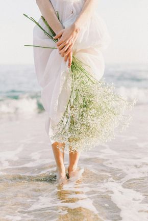 A woman in a white dress is holding a bouquet of baby 's breath on the beach.