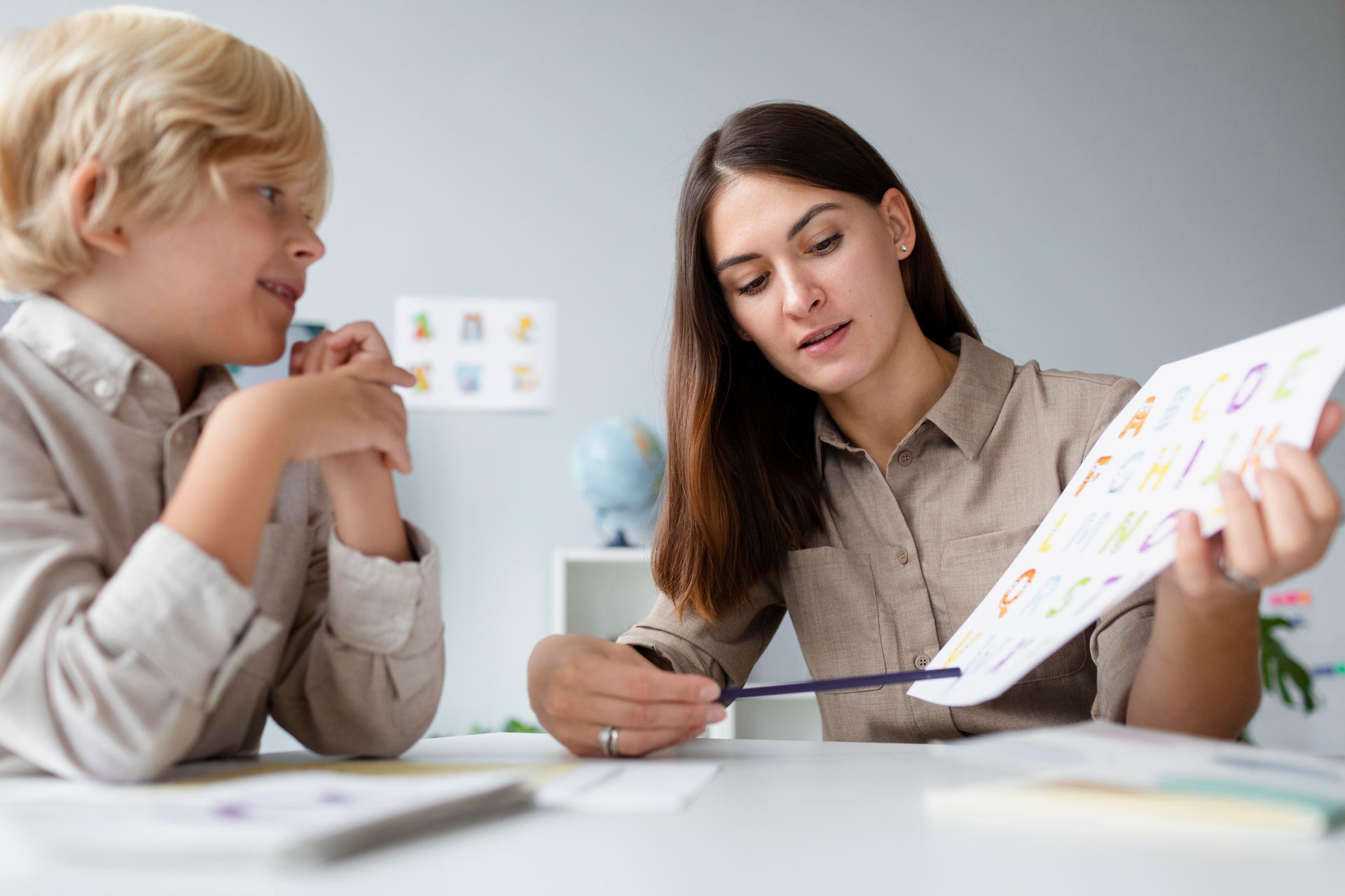 A woman points a pencil at a page while talking to a young boy, both seated at a table.