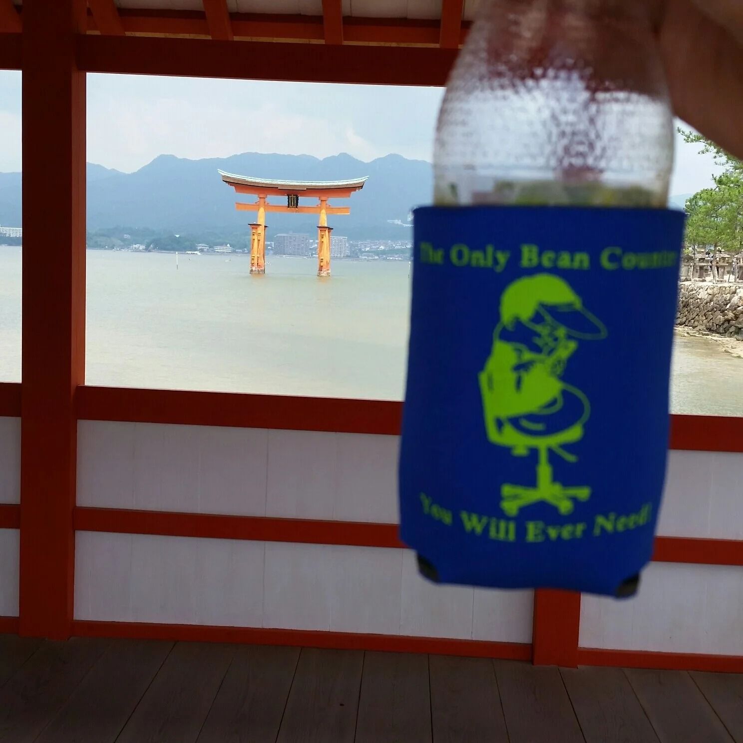 A drink in a blue koozie in front of a red torii gate in the water.