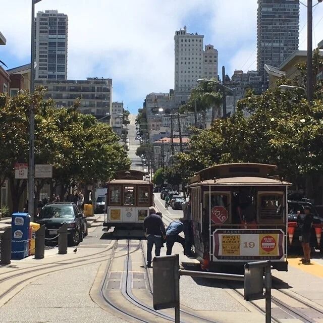 Cable cars on a steep San Francisco street. People stand nearby. Tall buildings and trees line the road.