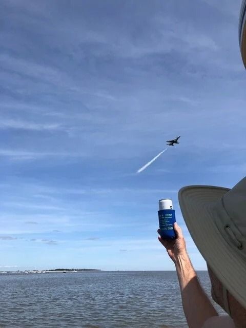 Person holding blue bottle watches a jet aircraft with smoke trail fly over water.