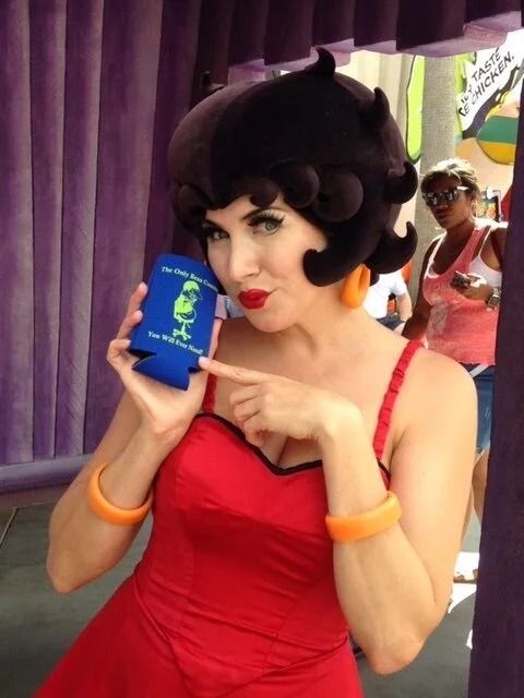 Woman dressed as Betty Boop, in a red dress, holding a blue drink holder and pointing.
