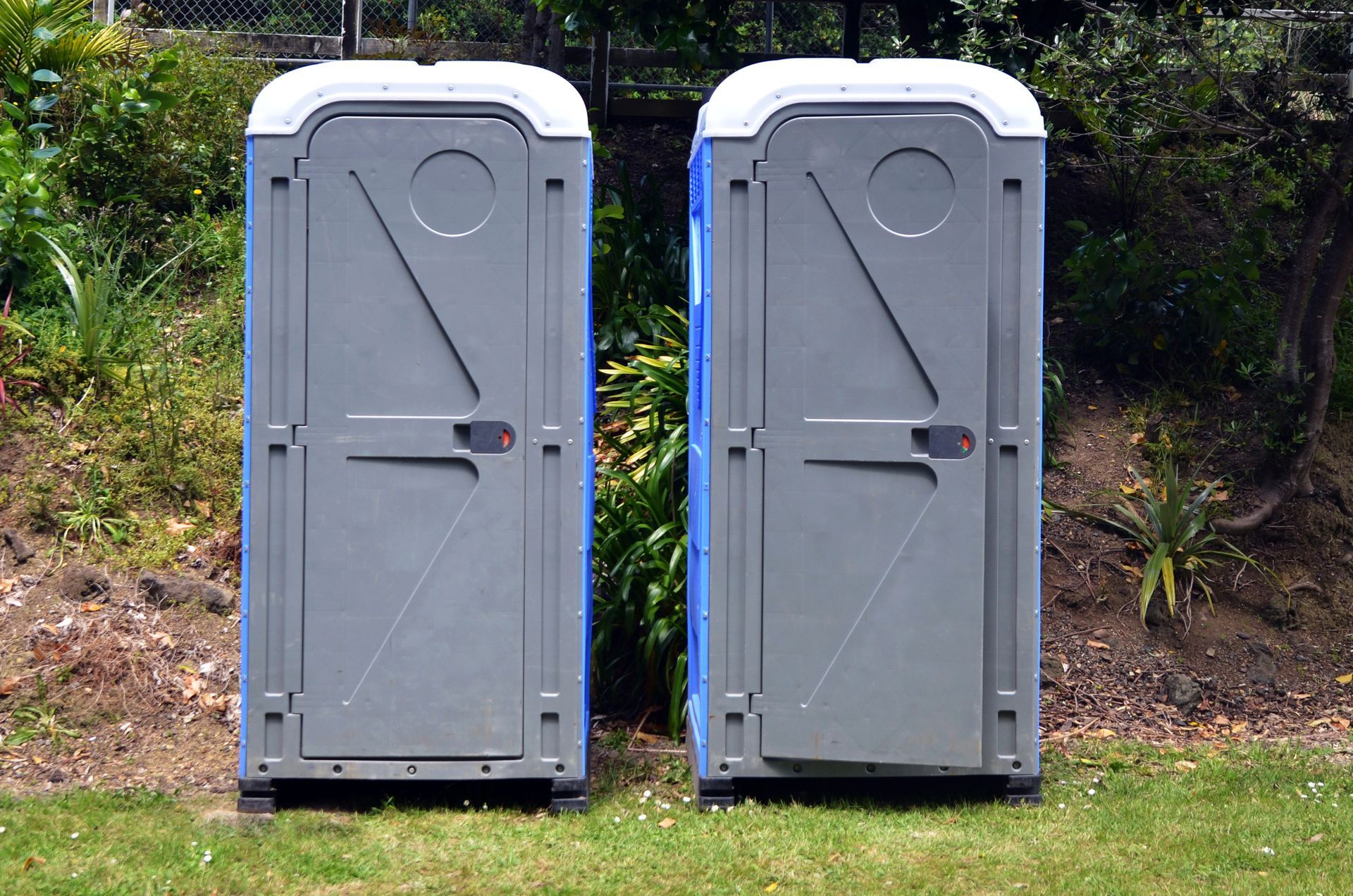 Dos baños portátiles grises sobre el césped con una valla y arbustos al fondo.