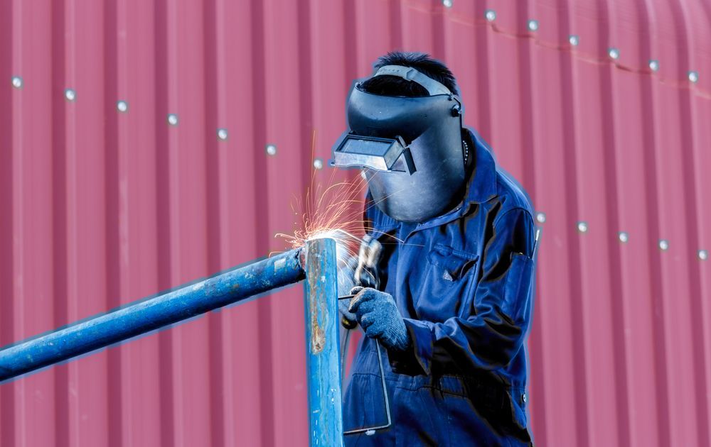 Welder in Blue Overalls Welding a Metal Pipe, Wearing a Protective Mask — GC Custom Fabrication in Tweed Heads, NSW