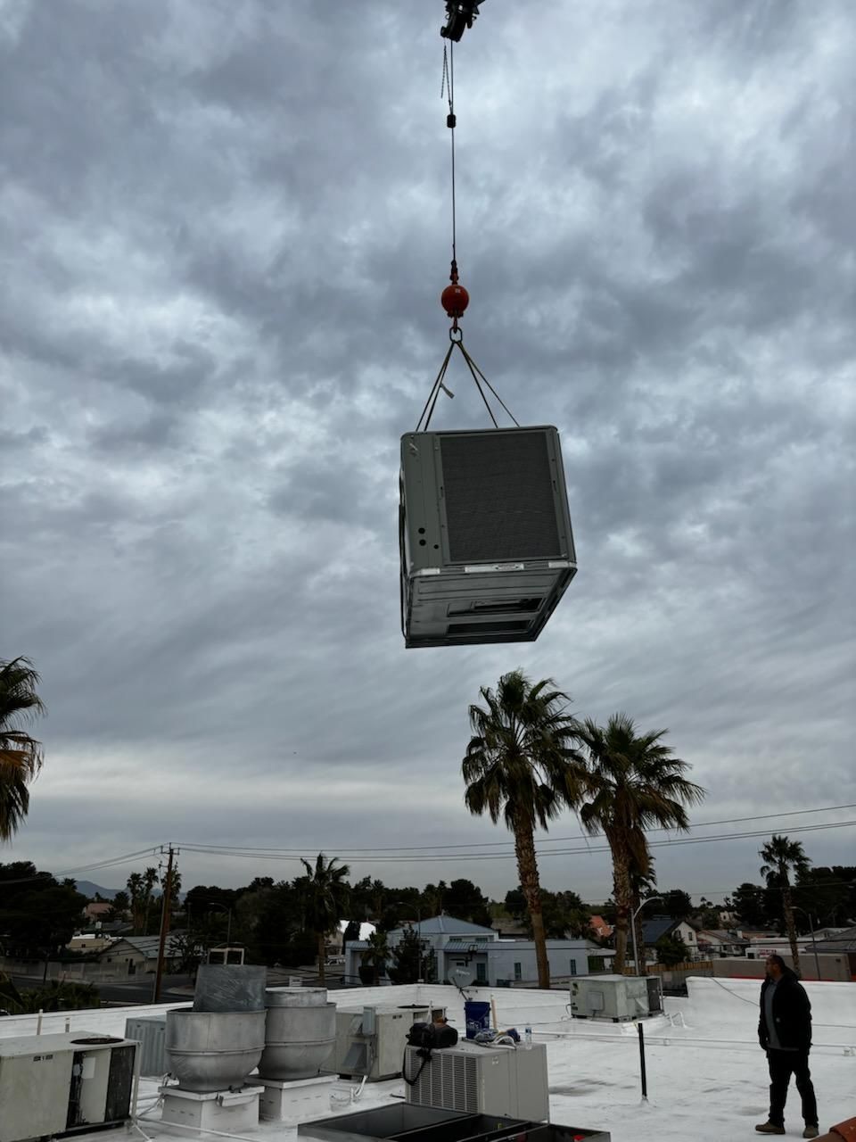 A crane lifts an HVAC unit onto a roof with palm trees under a cloudy sky.