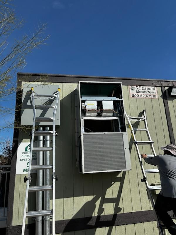 Workers install an HVAC unit on a building exterior with ladders under a blue sky.