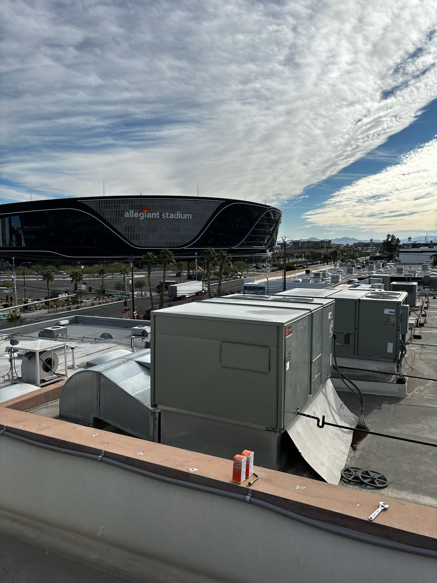A rooftop view of Allegiant Stadium, gray HVAC units, and a partly cloudy sky.
