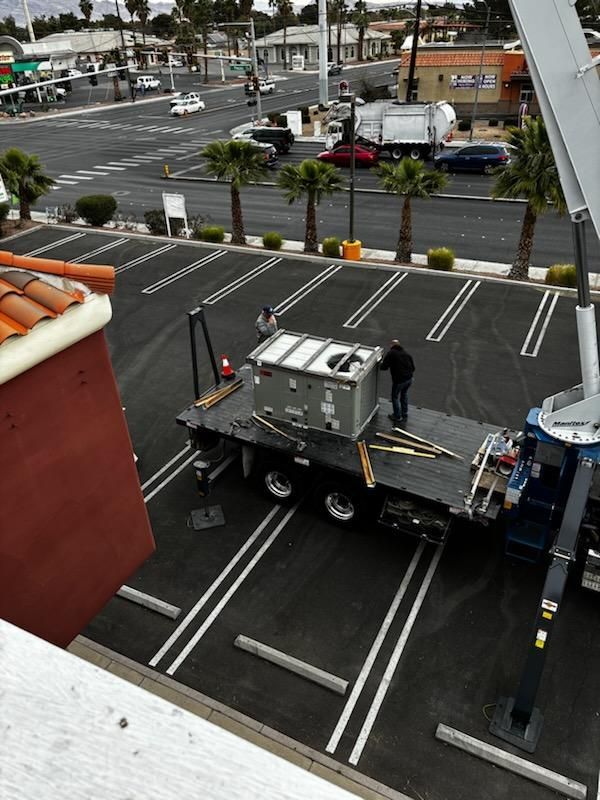 A large flatbed truck with a person secures a rectangular unit in a parking lot. A crane is extended overhead.