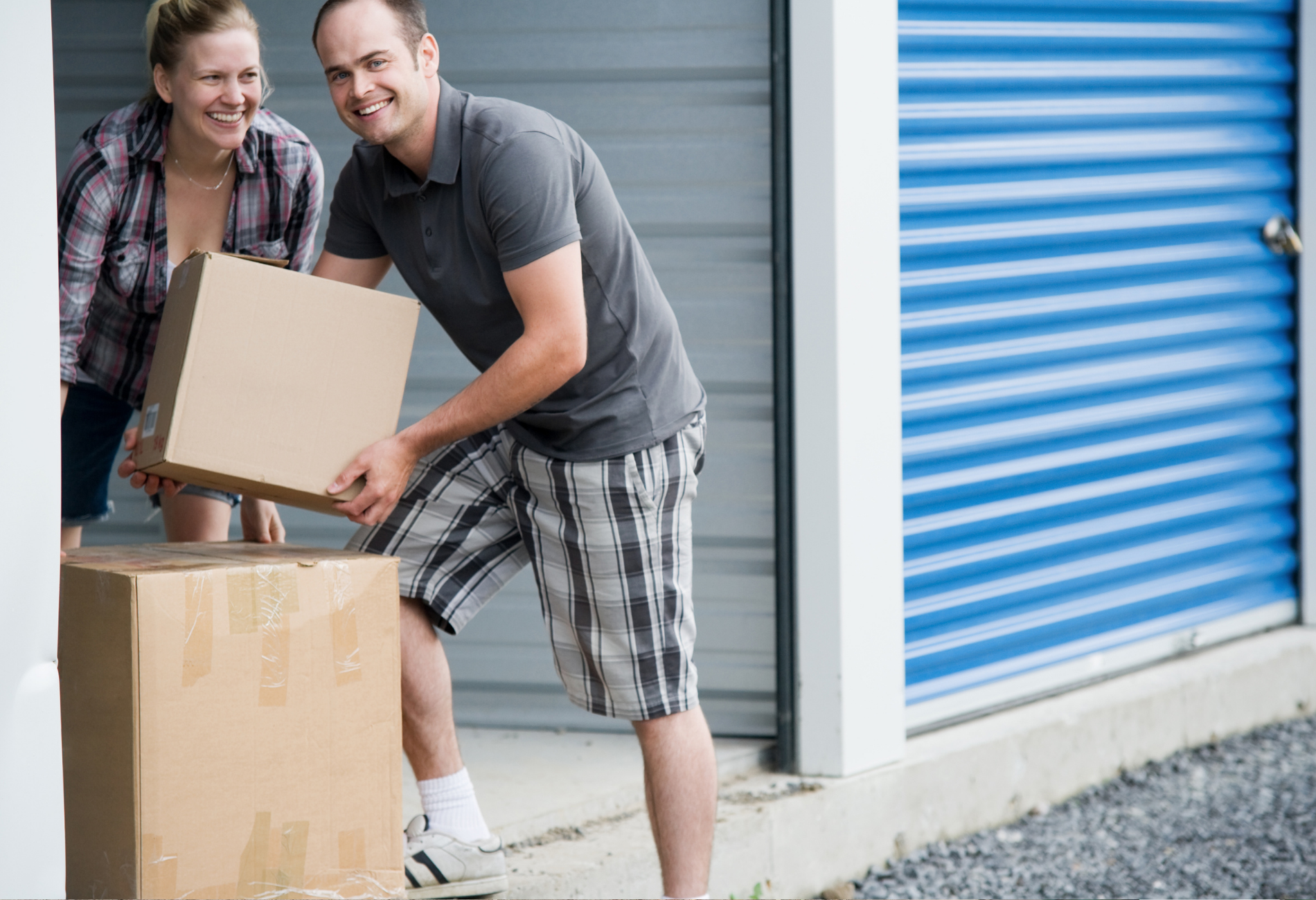 Couple unloading boxes at a storage unit, smiling. Gray and blue, concrete floor.
