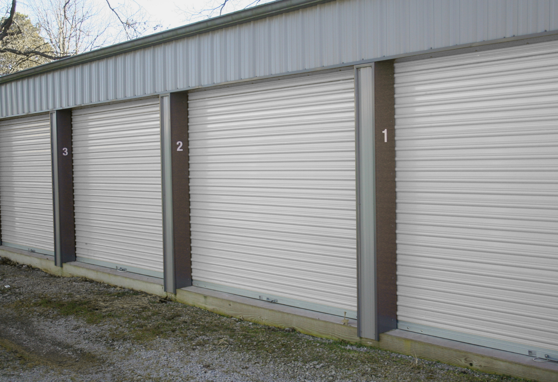 Storage units with roll-up doors, numbered 1, 2, and 3. Light gray metal siding and brown trim.