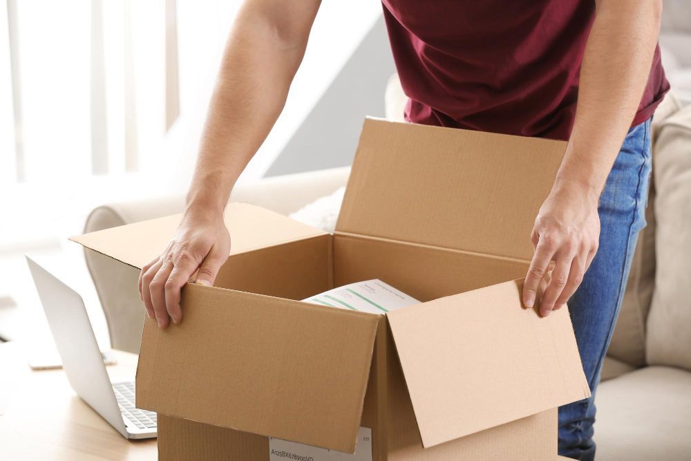 A man is opening a cardboard box with a laptop in the background.