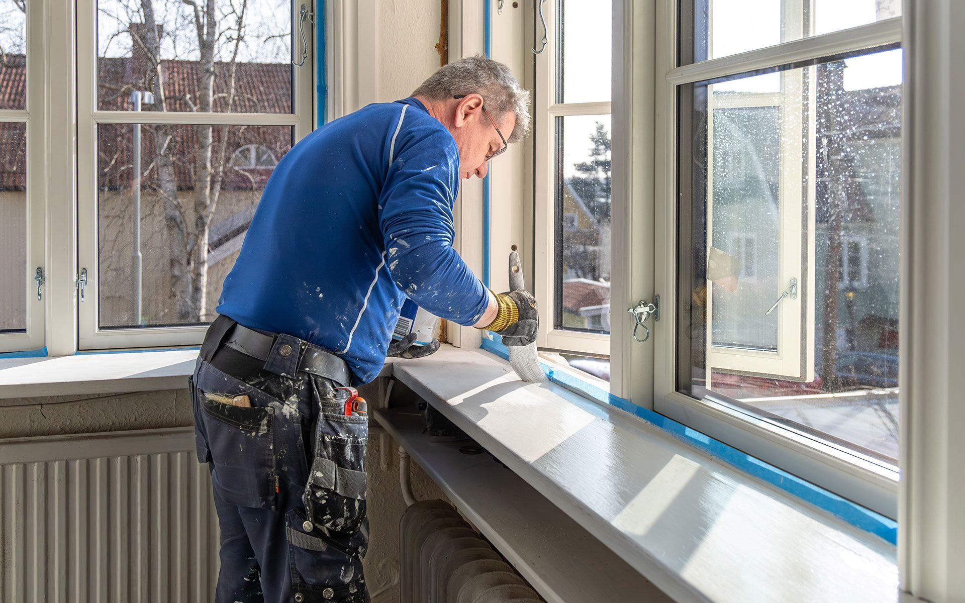 Man caulking a windowsill, near a window, wearing blue shirt and work pants.