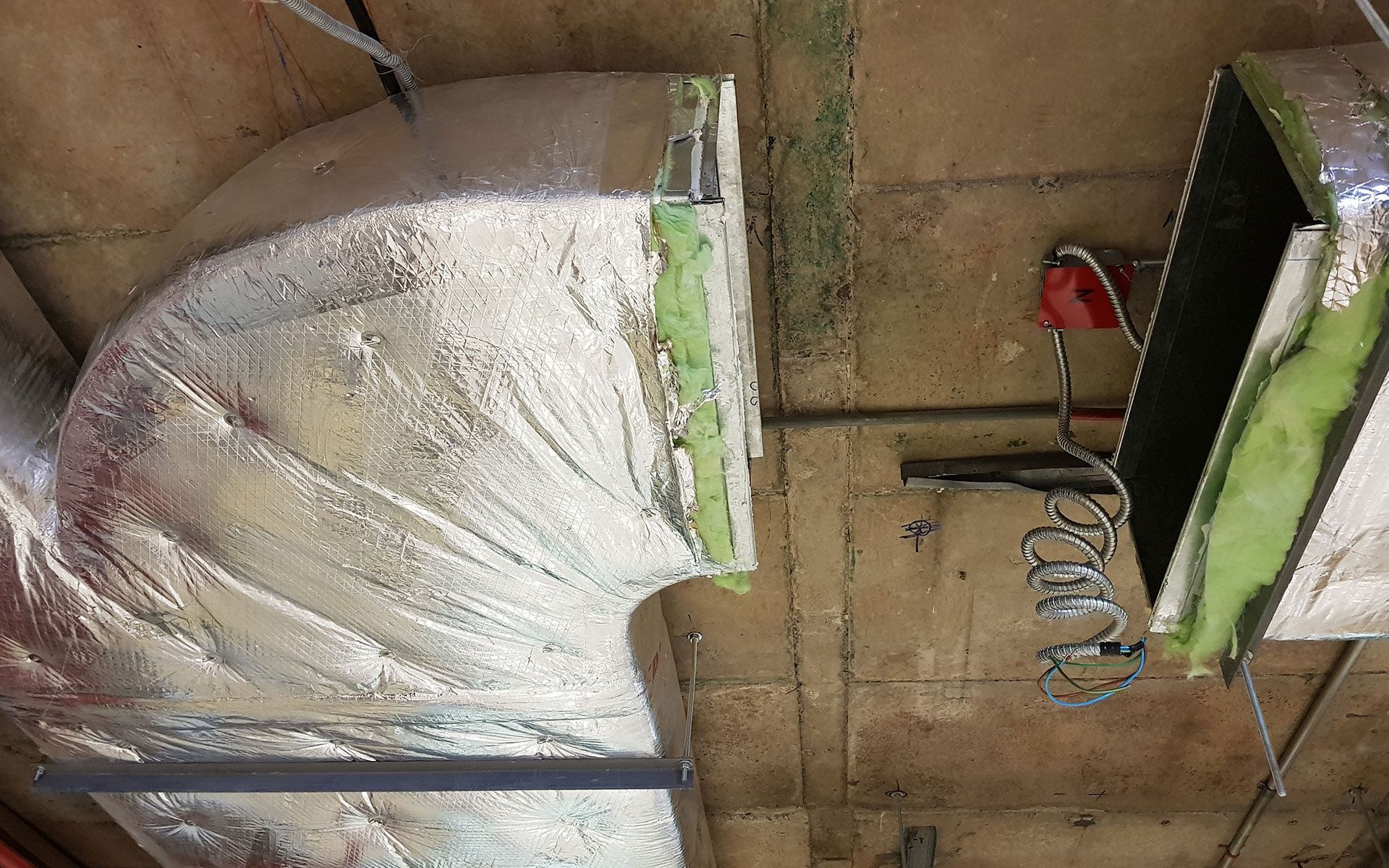 Insulated HVAC ductwork, silver wrap and green tape, mounted on a concrete ceiling.