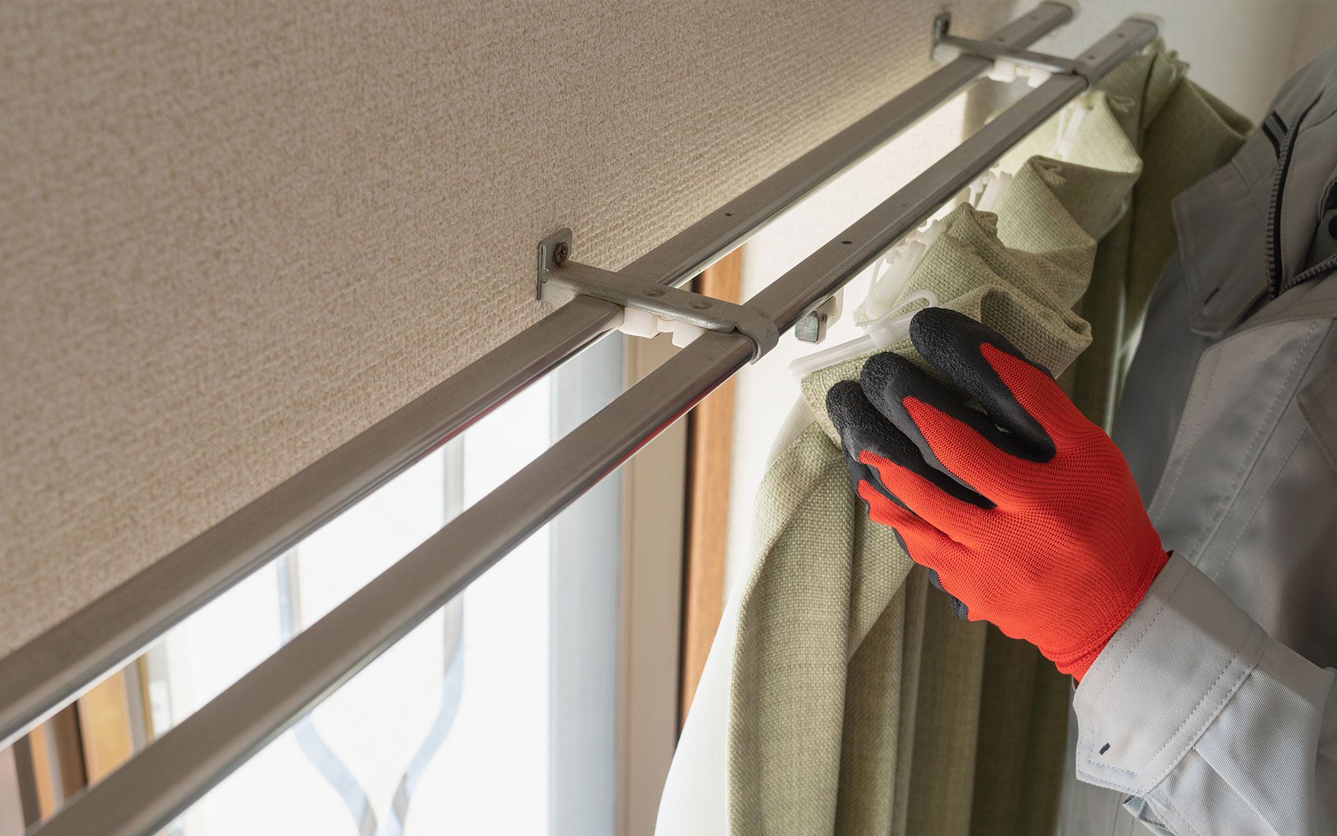Person wearing orange gloves adjusting a light-green curtain on a double curtain rod near a window.