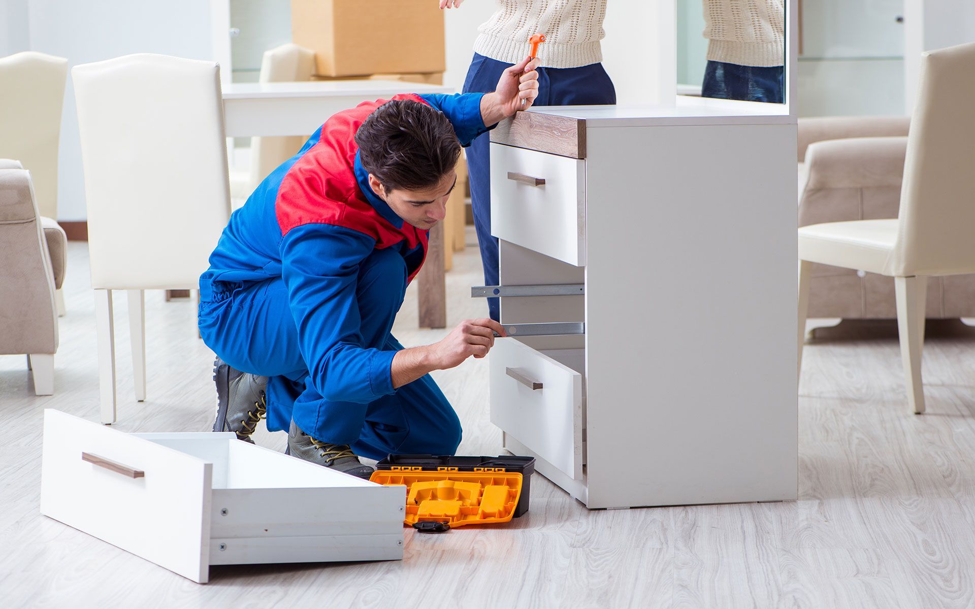 Man in blue coveralls assembling a white dresser with drawers, tools nearby, indoors.