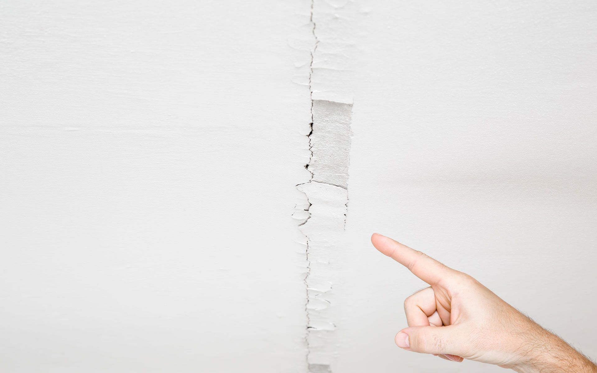 A hand points to a large crack in a white ceiling.
