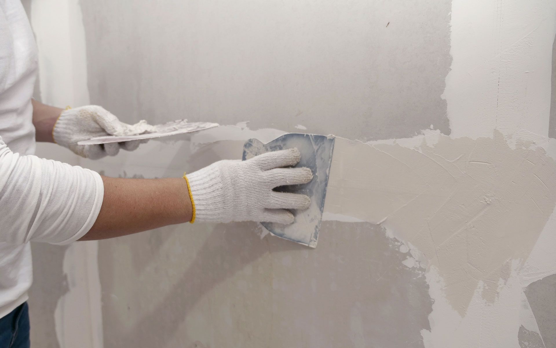 Person wearing gloves, using a putty knife to apply drywall compound to a wall.