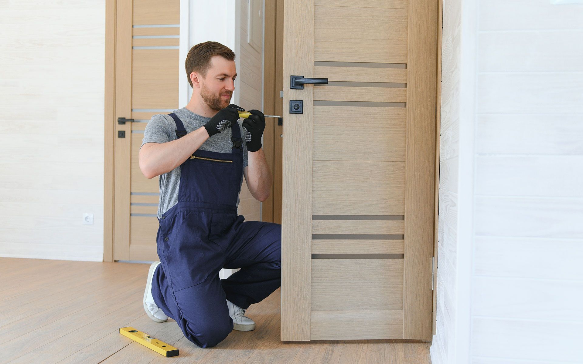 Man in blue overalls kneels, installing door hardware with a screwdriver. Wooden door and trim in a bright room.