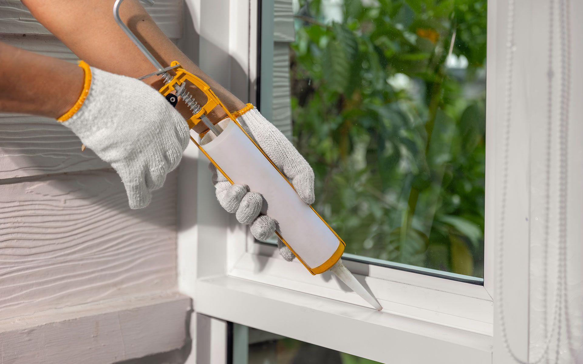 Person caulking around a window frame, wearing gloves, outdoors.