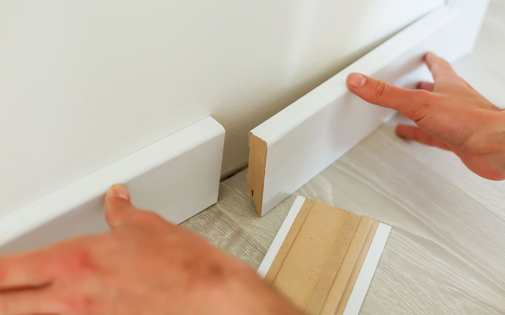 Hands installing white baseboard trim along a white wall.