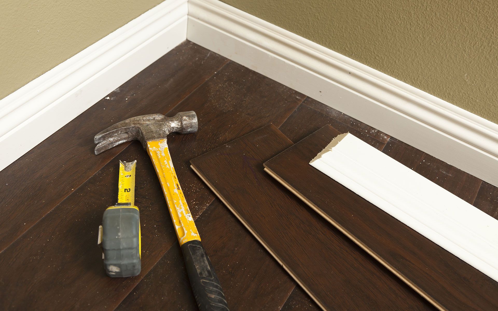 Hammer, tape measure, and flooring materials on dark hardwood floor next to white baseboard.