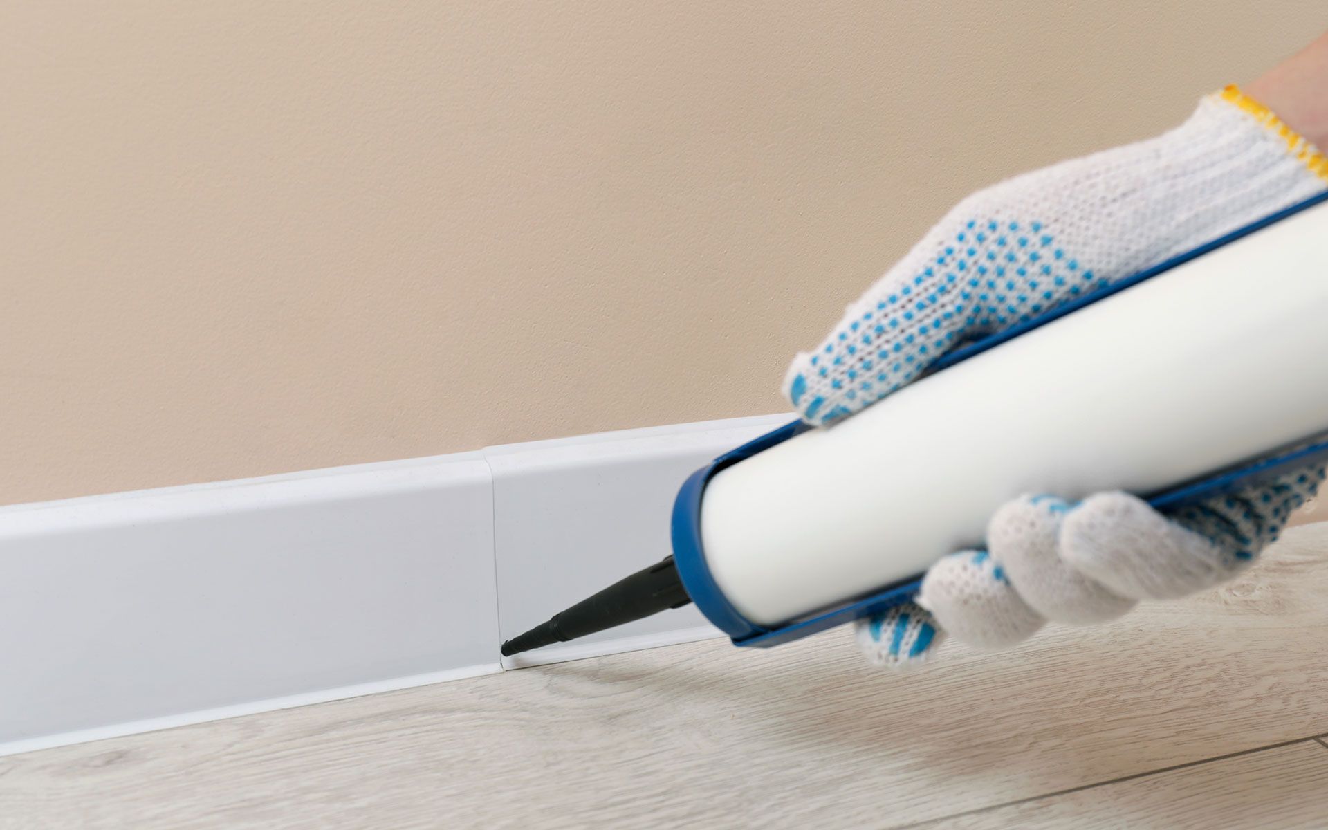 A person in a glove seals a white baseboard with caulk.