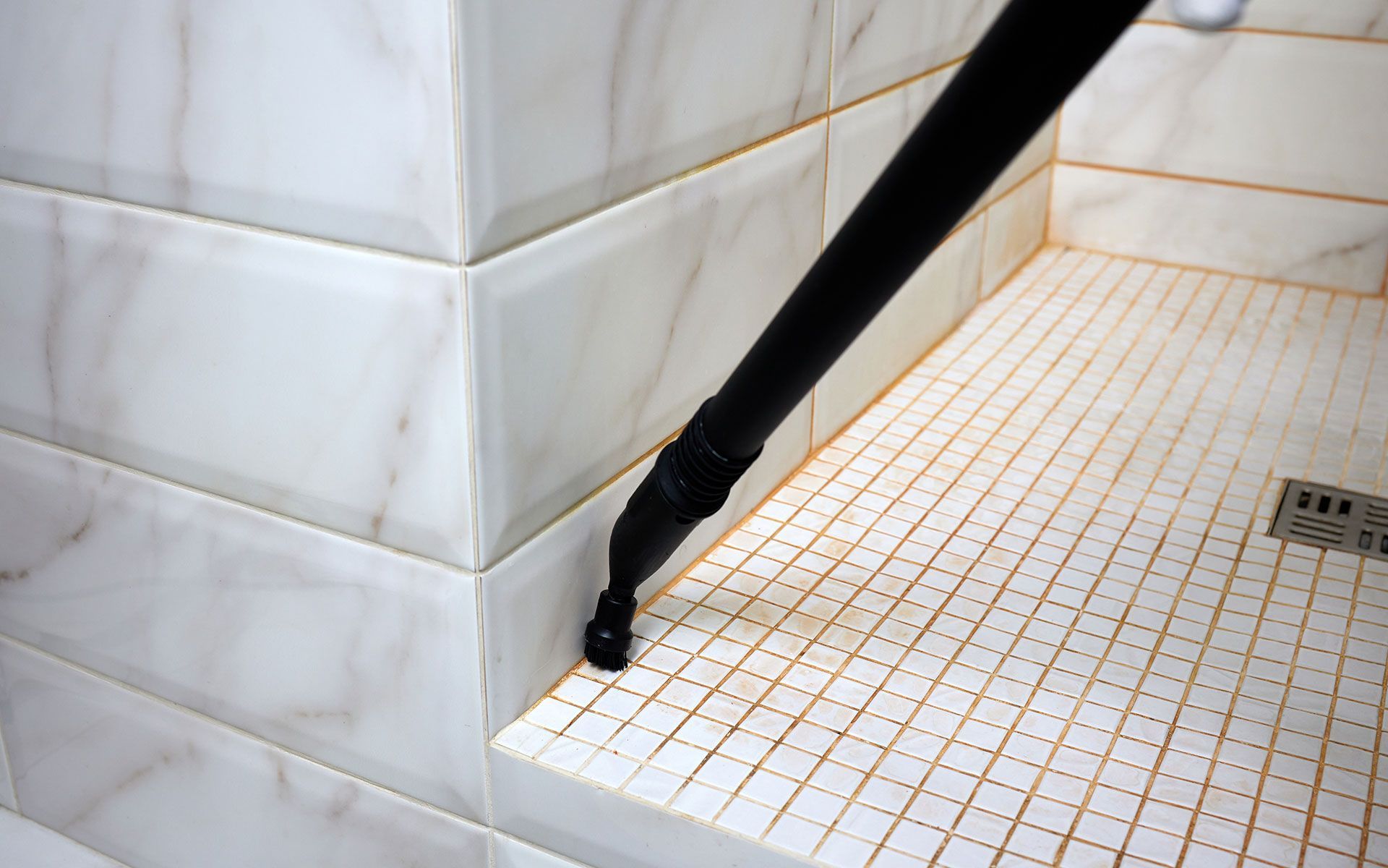 Black steam cleaner nozzle cleaning a white tiled shower floor.