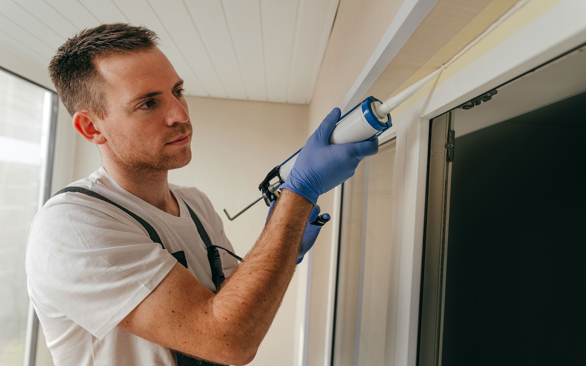 Man applying caulk around a window frame, wearing gloves and overalls, indoors.