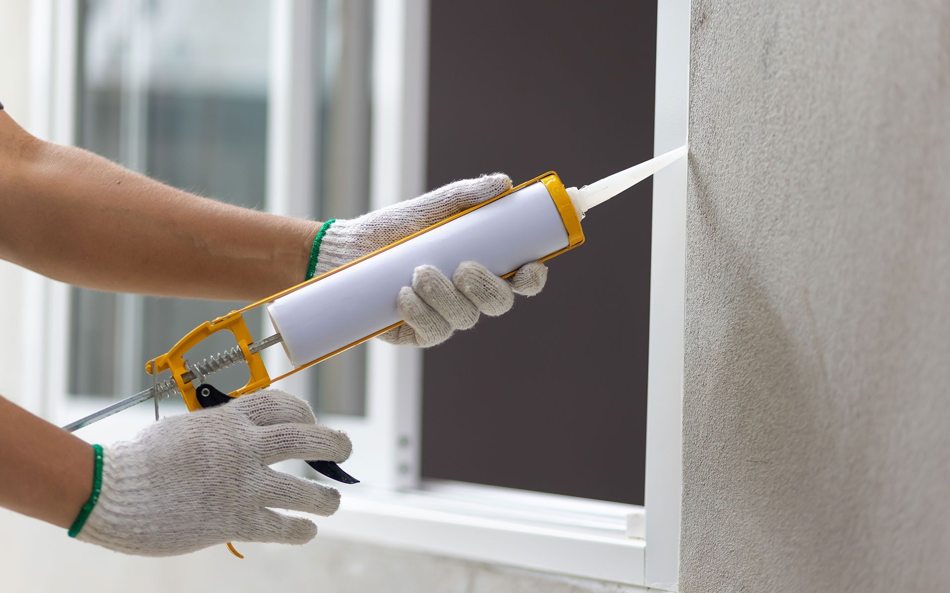 Person wearing gloves, caulking around a window frame with a caulk gun.