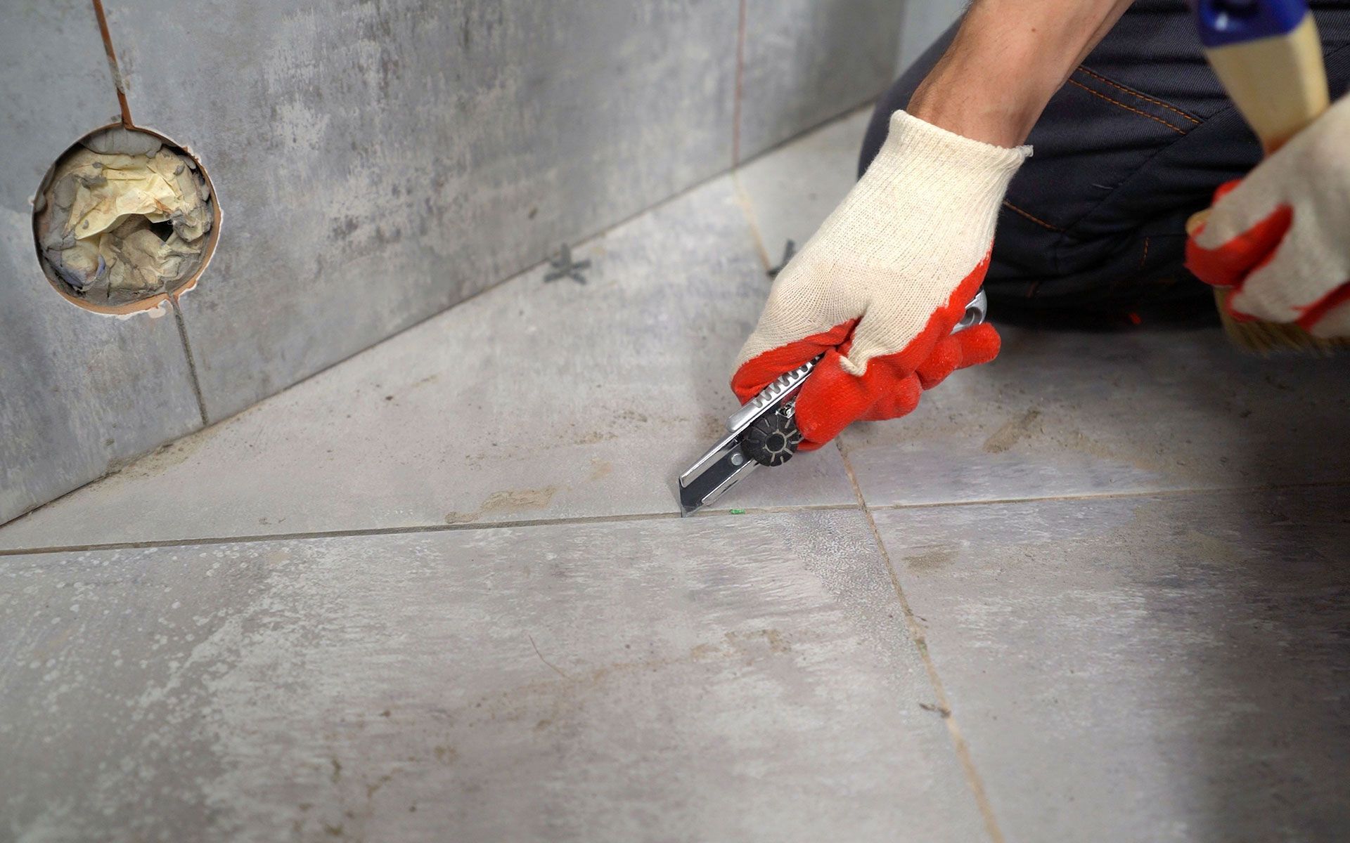 Person using a utility knife to cut grout between tiles on a bathroom floor.