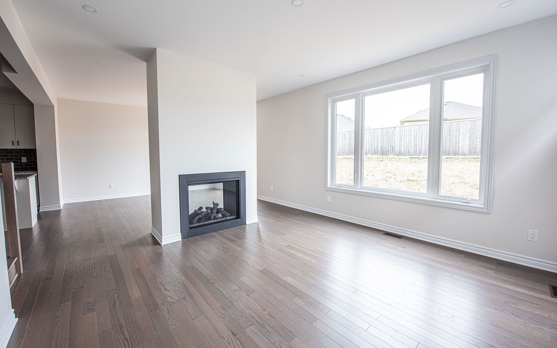 Empty living room with dark wood floor, fireplace, and large window overlooking a backyard.