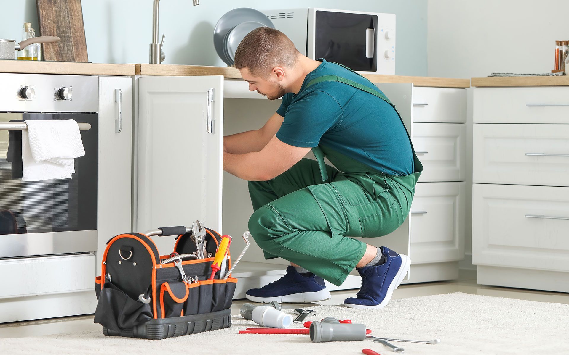 A person in green overalls crouches under a kitchen cabinet, working on plumbing, with tools and materials nearby.