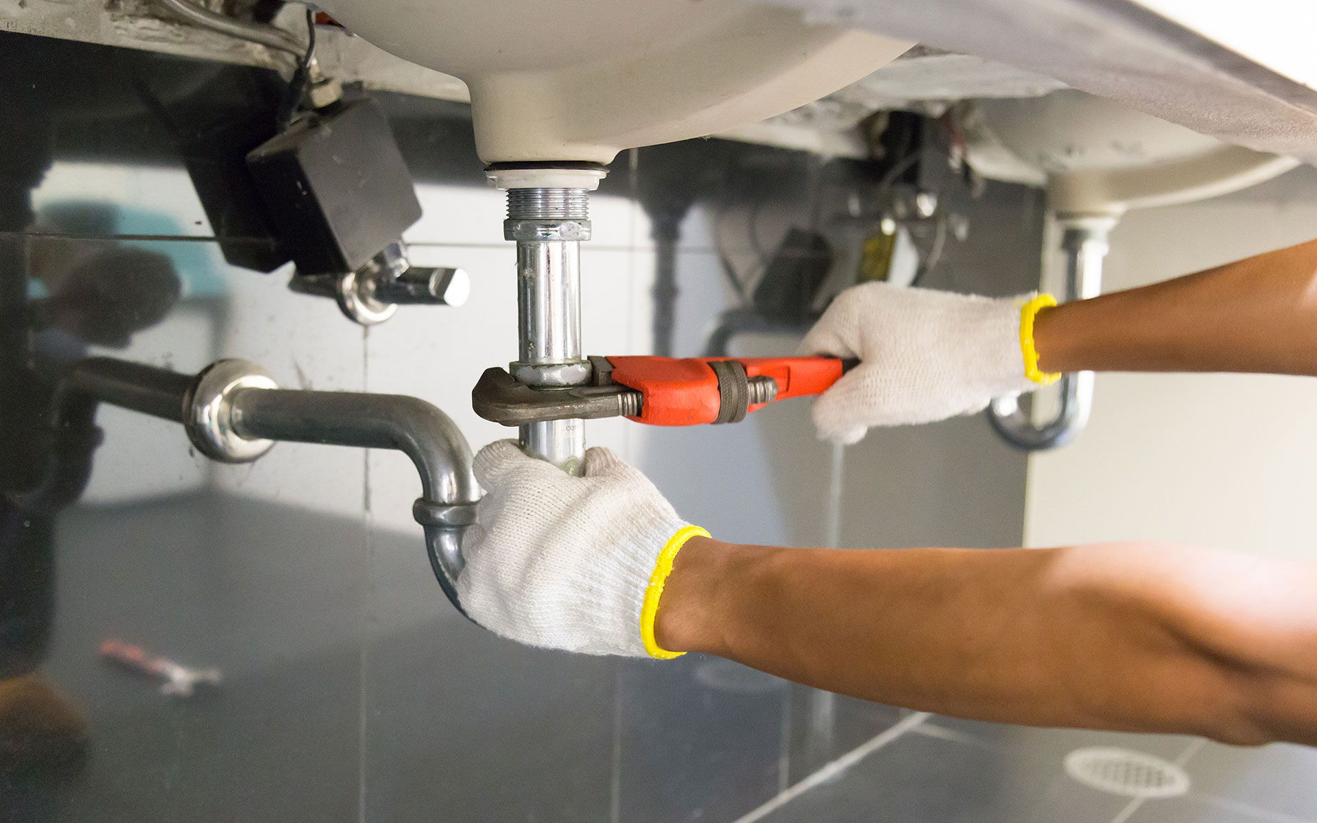 Hands wearing gloves tightening sink pipes with a wrench under a sink.