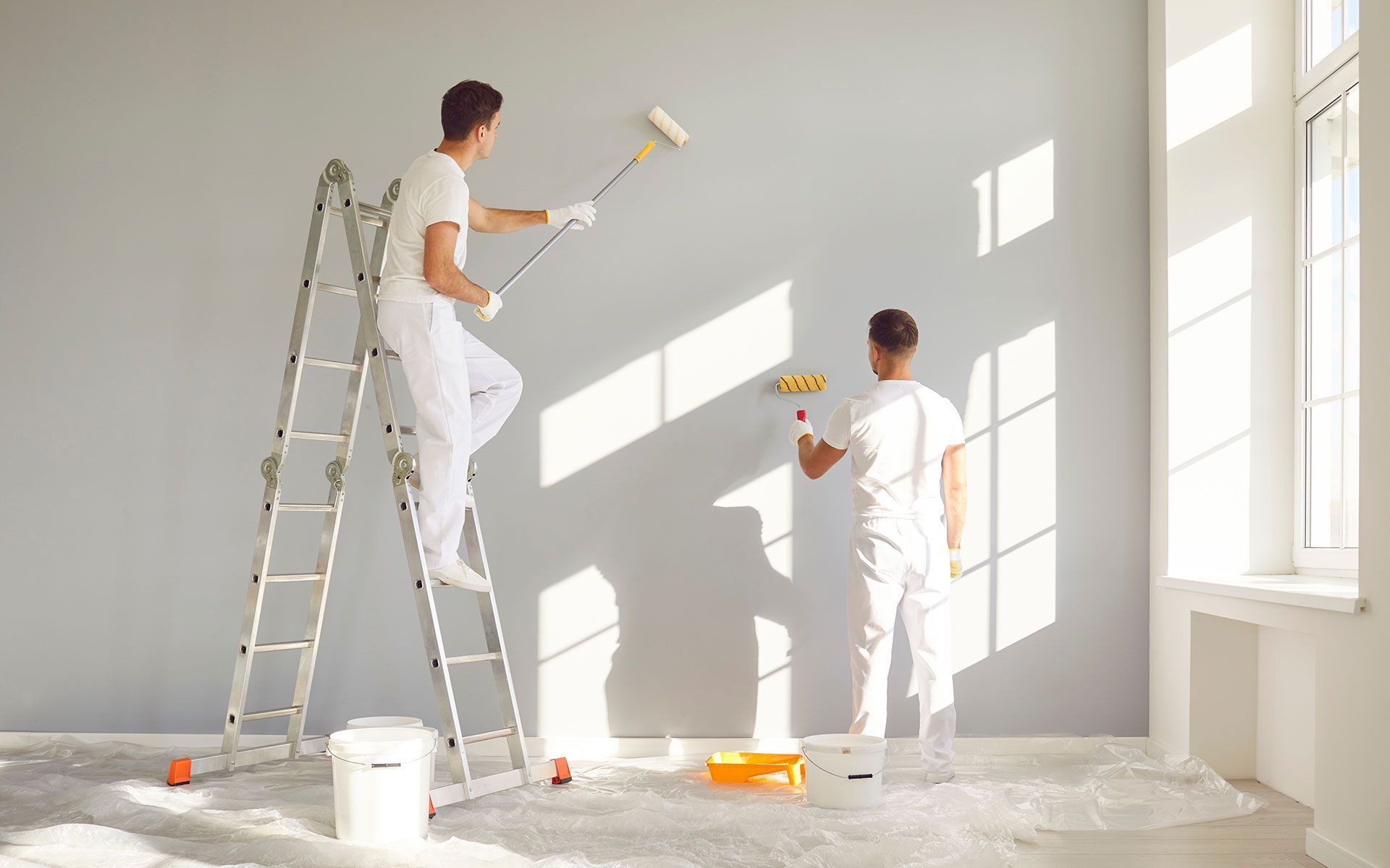 Two people painting a gray wall; one on a ladder using a roller, the other using a smaller roller.