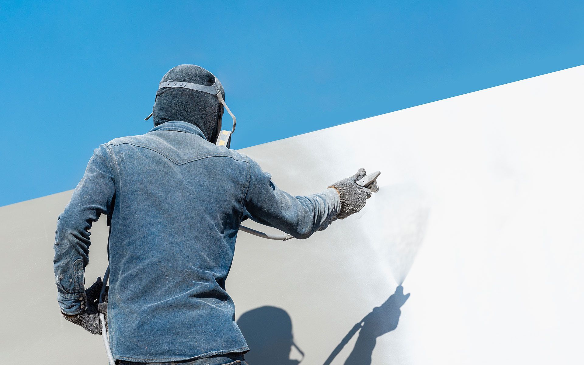 Person in protective gear sprays white paint on a wall outdoors under a blue sky.